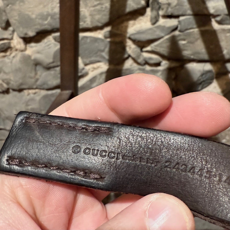 Gucci brown leather belt with silver-tone buckle, embossed branding, and adjustable strap, displayed on a wooden shelf.