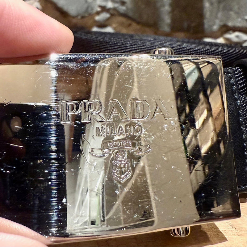 Prada black nylon belt with a silver-tone logo-engraved slide buckle, shown from various angles to highlight details such as the material, buckle engraving, and overall design.