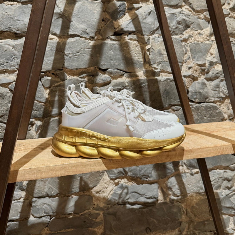 Versace White Gold Chain Reaction Sneakers featuring a white upper with translucent panels, textured fabric, embossed Greca patterns, and a bold gold chain-link sole. Includes Medusa head detailing on the heel and 'Chain Reaction' emblem on the tongue, with rope-style laces and pull tabs.