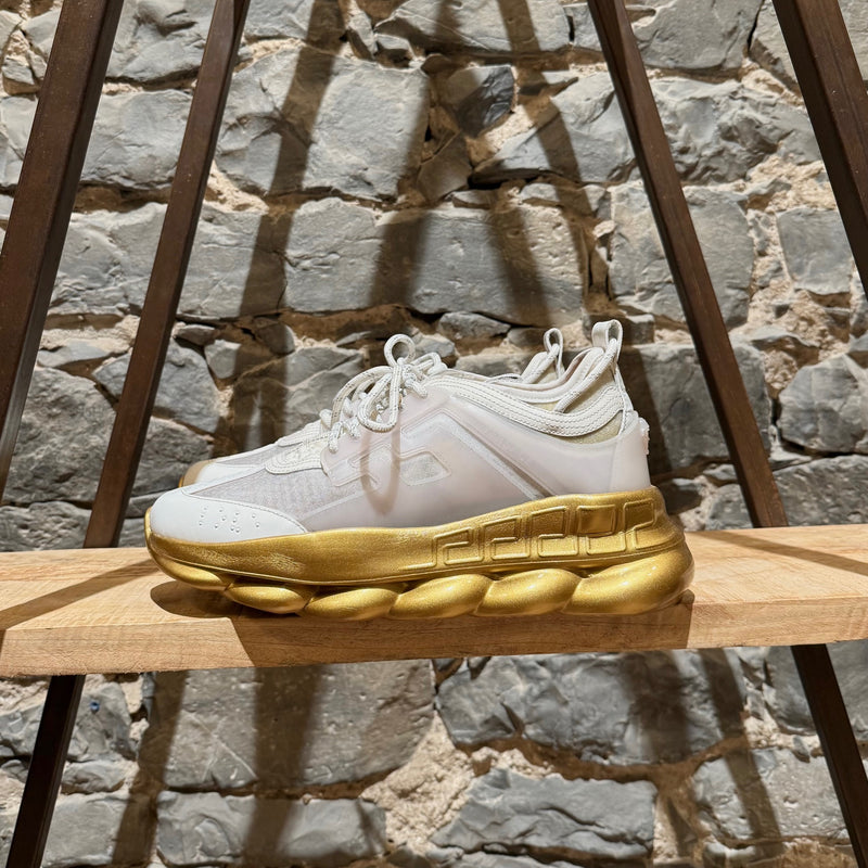 Versace White Gold Chain Reaction Sneakers featuring a white upper with translucent panels, textured fabric, embossed Greca patterns, and a bold gold chain-link sole. Includes Medusa head detailing on the heel and 'Chain Reaction' emblem on the tongue, with rope-style laces and pull tabs.