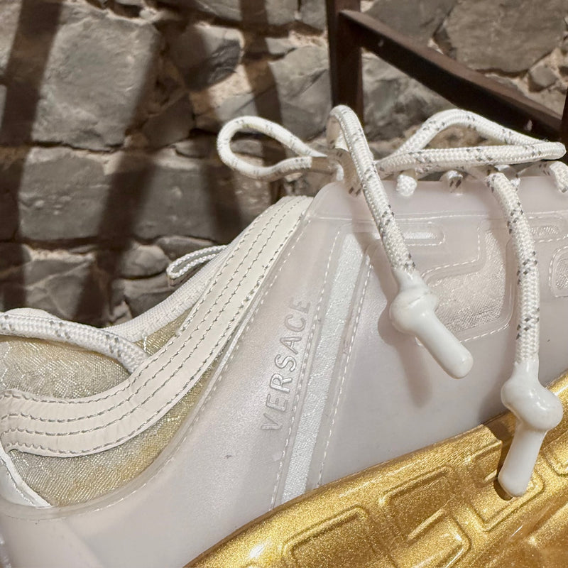 Versace White Gold Chain Reaction Sneakers featuring a white upper with translucent panels, textured fabric, embossed Greca patterns, and a bold gold chain-link sole. Includes Medusa head detailing on the heel and 'Chain Reaction' emblem on the tongue, with rope-style laces and pull tabs.