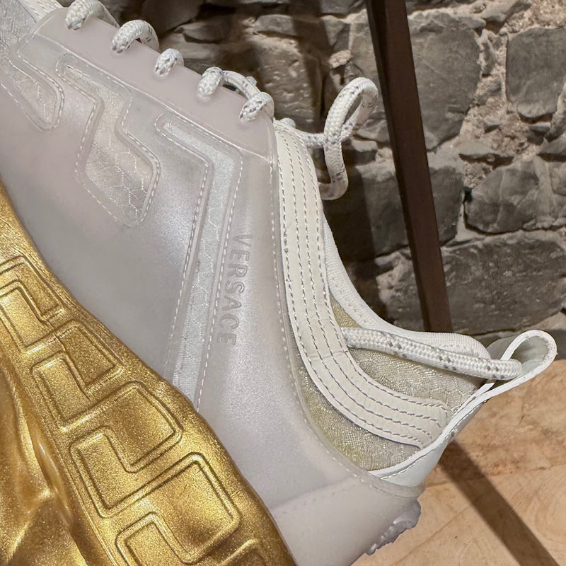 Versace White Gold Chain Reaction Sneakers featuring a white upper with translucent panels, textured fabric, embossed Greca patterns, and a bold gold chain-link sole. Includes Medusa head detailing on the heel and 'Chain Reaction' emblem on the tongue, with rope-style laces and pull tabs.